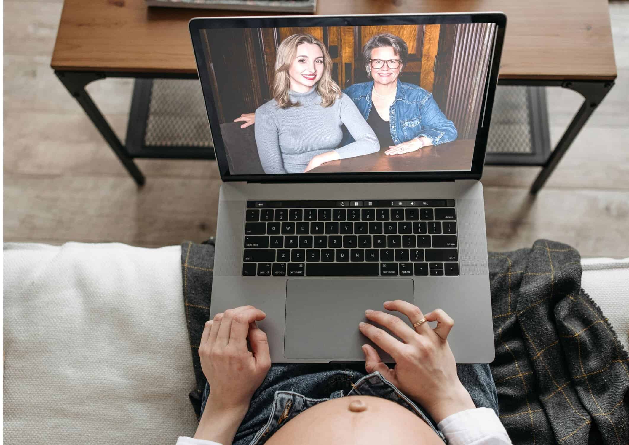 Trish and Chloe from Choice Surrogacy are sitting at a decorative table in front of a striped background and their image is shown on a a laptop placed on the lap of a pregnant woman. You can see the laptop and her belly, with Trish and Chloe on the screen, as they're having a virtual call.